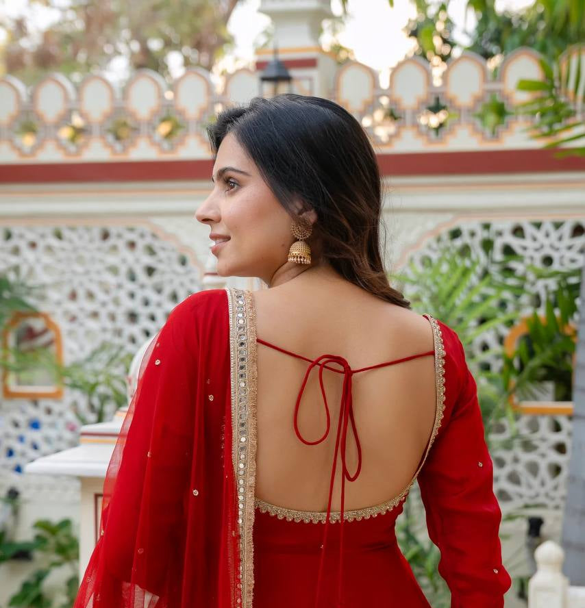 A woman wearing a red traditional dress with a deep open back, red tie straps, and a matching sheer dupatta with gold embroidery and tassel details, photographed outdoors in front of a decorative white railing and greenery.