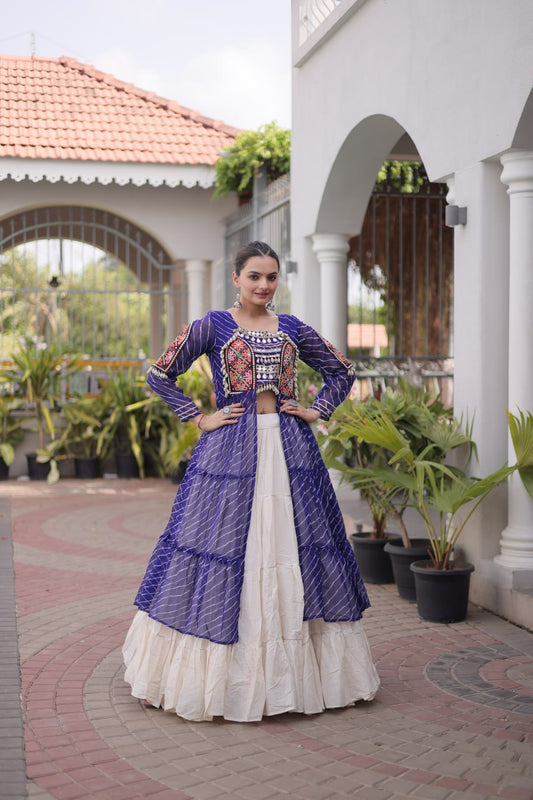 A woman stands outdoors in a courtyard wearing a traditional Indian outfit consisting of a purple embroidered kurta with a white skirt. The background features white walls with arches, potted plants, and a tiled roof.