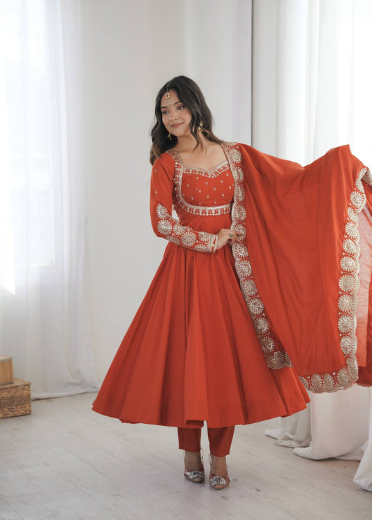 A woman modeling a traditional Indian outfit consisting of a rust-orange anarkali kurta with gold embroidery, matching pants, and a dupatta with ornate gold border detailing, set against a bright, minimalist studio background with white curtains.